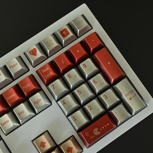 May include: A mechanical keyboard with a white frame and a mix of red and silver keycaps. The red keycaps are on the number pad and function keys, while the silver keys have white lettering. Some keys have symbols.