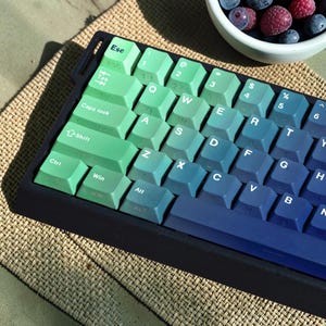 May include: A mechanical keyboard featuring a gradient colour scheme that transitions from light mint green on the left side to deep navy blue on the right side. The keycaps display white alphanumeric characters and function labels.