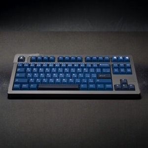 K&ouml;nnte beinhalten: Eine mechanische Tastatur mit einem dunkelgrauen Rahmen und marineblauen Tastenkappen. Die Tastatur hat ein Standardlayout mit alphanumerischen Tasten, Funktionstasten und einem Nummernblock. Die Tastenkappen zeigen sowohl englische Buchstaben als auch japanische Zeichen.