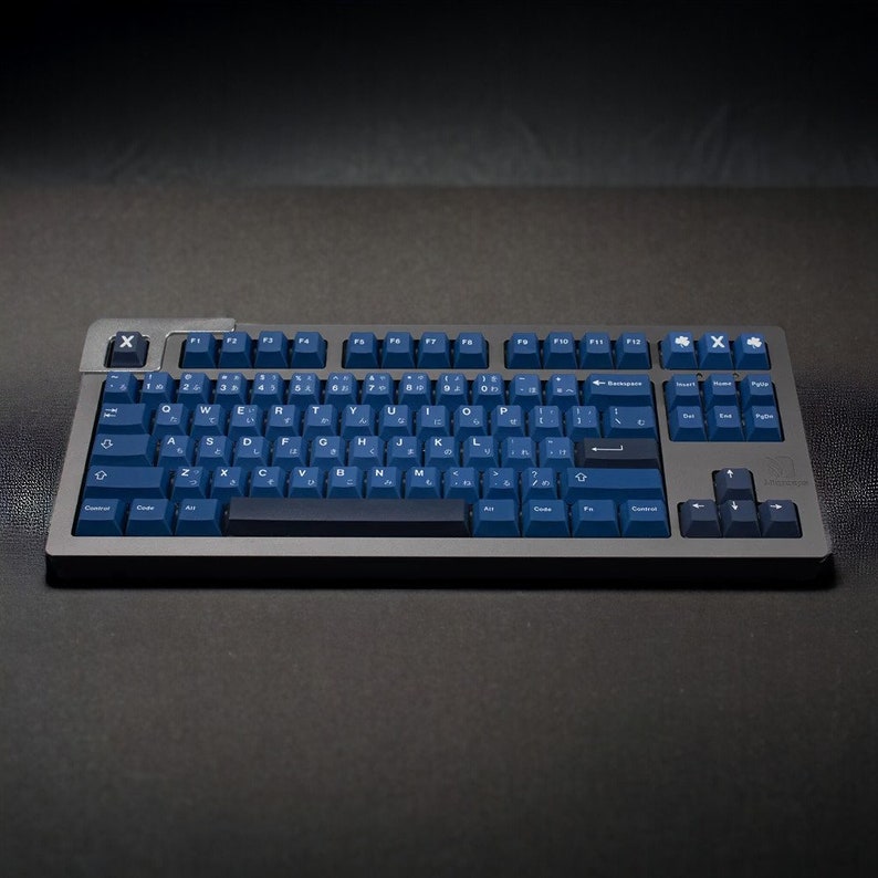 K&ouml;nnte beinhalten: Eine mechanische Tastatur mit einem dunkelgrauen Rahmen und marineblauen Tastenkappen. Die Tastatur hat ein Standardlayout mit alphanumerischen Tasten, Funktionstasten und einem Nummernblock. Die Tastenkappen zeigen sowohl englische Buchstaben als auch japanische Zeichen.