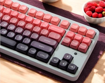 May include: A white and gray keyboard with pink and purple keys. The keyboard has a gradient color scheme, with the top row of keys being pink and the bottom row being purple. The keyboard has a variety of keys, including letters, numbers, symbols, and function keys. The keyboard is on a purple and gray surface.