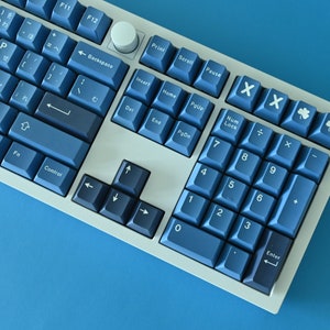 K&ouml;nnte beinhalten: Eine mechanische Tastatur mit blauen Tastenkappen und einem wei&szlig;en Rahmen. Die Tasten haben japanische Zeichen und englische Buchstaben. Der Ziffernblock und die Funktionstasten sind sichtbar. Die Tastatur liegt auf einer blauen Oberfl&auml;che.