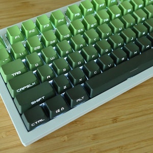 以下が含まれることがあります： 緑色のキーキャップが付いた白いメカニカルキーボード。キーキャップは、明るい緑色から暗い緑色へとグラデーションになっています。キーボードは、文字、数字、記号でラベル付けされたキーを持つ標準レイアウトです。キーボードは木製の表面にあります。