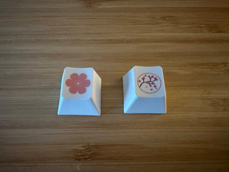 May include: Two white keycaps with pink floral designs. One keycap features a single pink flower, while the other has a pink cherry blossom branch.