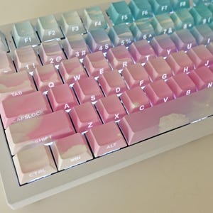 May include: A white mechanical keyboard with a gradient color scheme. The keys are pink, blue, and white, and the keyboard has a white frame. The keys are labeled with the standard keyboard layout, including the letters, numbers, and symbols.