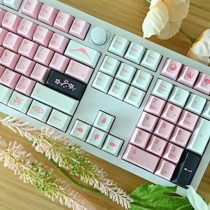 May include: A white mechanical keyboard with pink keycaps and Japanese-themed keycap designs. The keyboard features a white and pink colour scheme with a variety of keycap designs, including a mountain, a cherry blossom branch, a torii gate, and a fan.