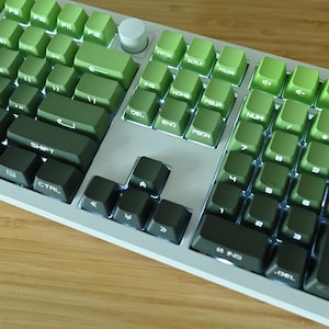 以下が含まれることがあります： 緑色のキーキャップが付いた白いメカニカルキーボード。キーキャップは明るい緑色から暗い緑色へとグラデーションになっています。キーボードは、ファンクションキー、矢印キー、テンキーを備えた標準レイアウトです。
