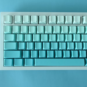 Può includere: Una tastiera meccanica bianca con tasti a gradiente blu e bianco. La tastiera ha un layout standard con tasti funzione, tasti numerici e una barra spaziatrice.