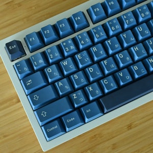 K&ouml;nnte beinhalten: Mechanische Tastatur mit blauen Tasten und wei&szlig;em Rahmen auf einer Holzoberfl&auml;che. Die Tasten zeigen wei&szlig;e japanische Zeichen und englische Buchstaben. Die Tastatur hat Funktionstasten und eine Steuerungstaste.