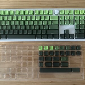 以下が含まれることがあります： メカニカルキーボード用の緑と黒のキーキャップセット。キーキャップは透明なプラスチックカバー付きのトレイに配置されています。キーキャップは緑色のグラデーションで、一番暗い緑が下、一番明るい緑が上になっています。