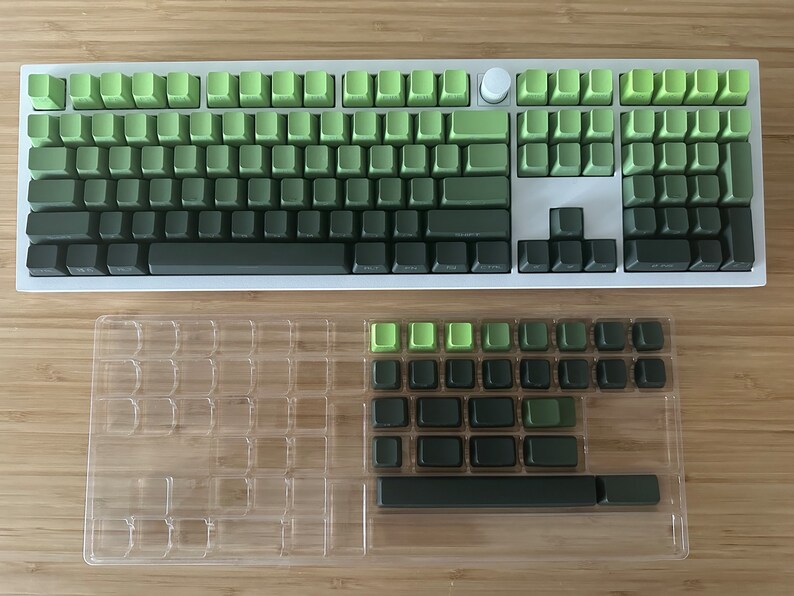 以下が含まれることがあります： メカニカルキーボード用の緑と黒のキーキャップセット。キーキャップは透明なプラスチックカバー付きのトレイに配置されています。キーキャップは緑色のグラデーションで、一番暗い緑が下、一番明るい緑が上になっています。