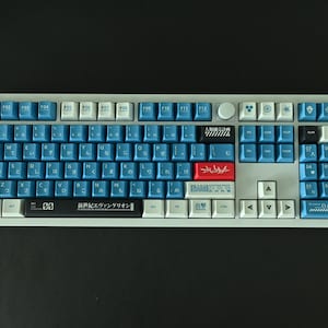 Op de afbeelding: Een wit en grijs mechanisch toetsenbord met blauwe en rode toetsen. Het toetsenbord heeft een Japanse lay-out met de tekst "新世紀エヴァンゲリオン" gedrukt op de spatiebalk.