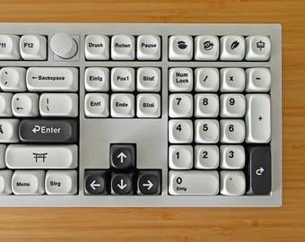 Peut inclure: Un clavier blanc avec des touches noires, comportant divers symboles et lettres. Le clavier possède un pavé numérique à droite et une rangée de touches de fonction en haut. Les touches sont étiquetées en allemand.