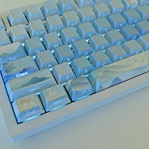 Könnte beinhalten: Eine weiße mechanische Tastatur mit blauer Hintergrundbeleuchtung. Die Tasten haben ein hellblaues und weißes Bergmotiv. Die Tasten sind mit dem Standard-Tastaturlayout beschriftet, einschließlich Esc, Tab, Caps Lock, Shift, Ctrl, Win, F1-F6, 1-0, Q-Z und A-Z.