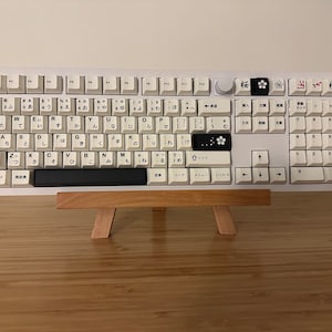 K&ouml;nnte beinhalten: Eine wei&szlig;e mechanische Tastatur mit schwarzen Tastenkappen, die japanische Zeichen und Symbole zeigen. Die Tastatur steht auf einem Holzst&auml;nder vor einem wei&szlig;en Hintergrund.