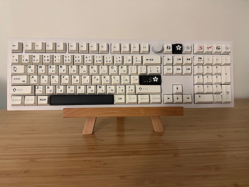 K&ouml;nnte beinhalten: Eine wei&szlig;e mechanische Tastatur mit schwarzen Tastenkappen, die japanische Zeichen und Symbole zeigen. Die Tastatur steht auf einem Holzst&auml;nder vor einem wei&szlig;en Hintergrund.