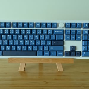 K&ouml;nnte beinhalten: Eine mechanische Tastatur in voller Gr&ouml;&szlig;e mit wei&szlig;em Rahmen und dunkelblauen Tastenkappen. Die Tasten verf&uuml;gen &uuml;ber japanische Zeichen und standardm&auml;&szlig;ige englische Buchstaben. Die Tastatur wird auf einer kleinen Holzstaffelei pr&auml;sentiert, die ihr Design und ihre Funktionalit&auml;t hervorhebt.
