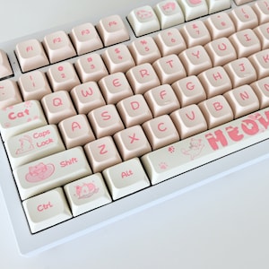 Pode incluir: Um teclado branco com teclas rosa com designs temáticos de gatos. O teclado tem uma tecla rosa "MEOW" na barra de espaço.