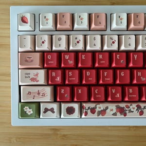 May include: A mechanical keyboard featuring a white frame and a custom keycap set in shades of red, pink, and cream. The keys display various illustrations including strawberries, flowers, musical notes, and small animals, alongside standard QWERTY lettering.
