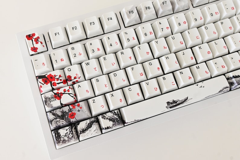 May include: A white mechanical keyboard with Japanese keycaps and a custom design featuring black and white ink drawings of cherry blossoms, mountains, and a boat. The keycaps have red Japanese characters.