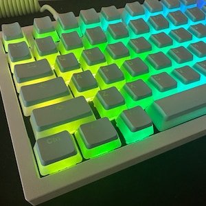 May include: A white mechanical keyboard with rainbow backlit keys. The keys are labeled with the standard QWERTY layout.