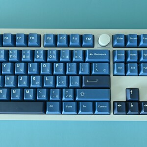 K&ouml;nnte beinhalten: Eine mechanische Tastatur mit blauen Tastenkappen und einem wei&szlig;en Rahmen. Die Tasten sind in einem Standard-Layout angeordnet, mit japanischen Zeichen auf einigen Tasten. Ein wei&szlig;er Drehknopf ist vorhanden.