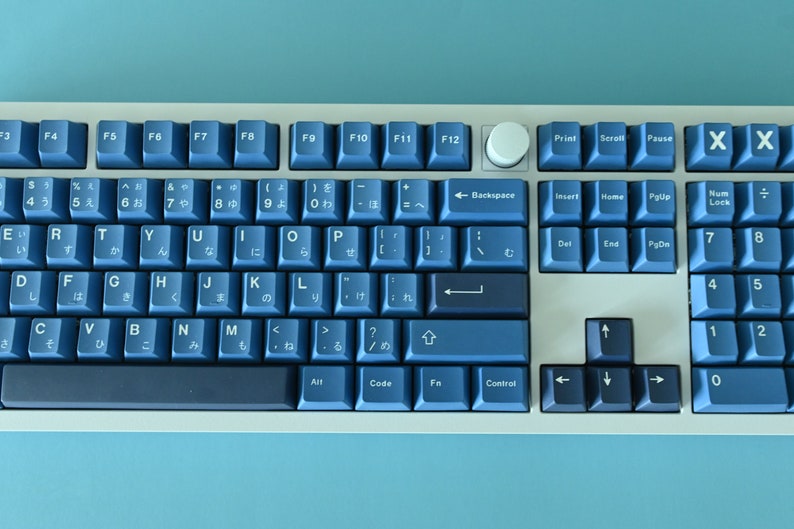 K&ouml;nnte beinhalten: Eine mechanische Tastatur mit blauen Tastenkappen und einem wei&szlig;en Rahmen. Die Tasten sind in einem Standard-Layout angeordnet, mit japanischen Zeichen auf einigen Tasten. Ein wei&szlig;er Drehknopf ist vorhanden.