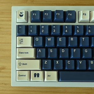May include: A mechanical keyboard with white and navy blue keycaps. The keyboard has a light grey frame and is set against a wooden surface. The keycaps feature various symbols and letters.