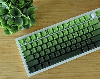 May include: A white mechanical keyboard with green and dark green keycaps. The keycaps are a gradient from light green to dark green. The keyboard is on a wooden surface.