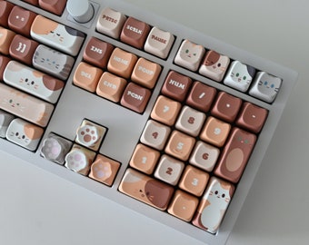 Könnte beinhalten: Eine mechanische Tastatur mit individuellen Tastenkappen im Katzen-Design. Die Tastenkappen zeigen verschiedene Katzenillustrationen und Pfotenabdrücke in Braun-, Weiß- und Pfirsichtönen. Die Tastatur hat einen weißen Rahmen und einen Lautstärkeregler.