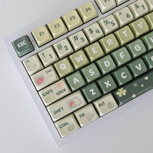 May include: A white mechanical keyboard with green and beige keys. The keys have various symbols and characters, including the letters of the alphabet, numbers, and punctuation marks. The keyboard has a white frame and a white base. The keys have a light green and beige color scheme, with some keys having a darker green color. The keys have a variety of symbols and characters, including the letters of the alphabet, numbers, and punctuation marks. The keyboard has a white frame and a white base.