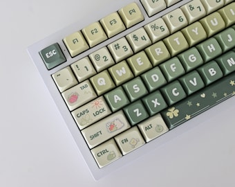 May include: A white mechanical keyboard with green and beige keys. The keys have various symbols and characters, including the letters of the alphabet, numbers, and punctuation marks. The keyboard has a white frame and a white base. The keys have a light green and beige color scheme, with some keys having a darker green color. The keys have a variety of symbols and characters, including the letters of the alphabet, numbers, and punctuation marks. The keyboard has a white frame and a white base.