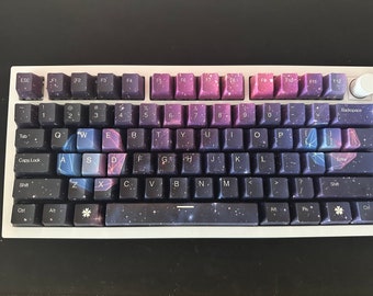 May include: A keyboard with a galaxy-themed design. The keys are black with a white starry pattern and the top row of function keys are purple with white lettering. The keyboard has a white frame and a white space bar.
