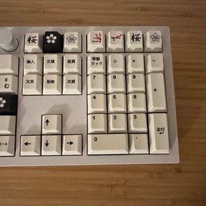 K&ouml;nnte beinhalten: Eine wei&szlig;e Tastatur mit schwarzen Tasten und japanischen Schriftzeichen. Die Tasten haben ein florales Design mit Kirschbl&uuml;ten und Zweigen. Die Tastatur verf&uuml;gt &uuml;ber einen Nummernblock und Pfeiltasten.