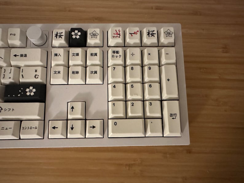 K&ouml;nnte beinhalten: Eine wei&szlig;e Tastatur mit schwarzen Tasten und japanischen Schriftzeichen. Die Tasten haben ein florales Design mit Kirschbl&uuml;ten und Zweigen. Die Tastatur verf&uuml;gt &uuml;ber einen Nummernblock und Pfeiltasten.