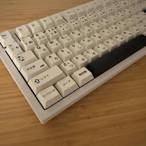 K&ouml;nnte beinhalten: Eine wei&szlig;e mechanische Tastatur mit japanischen Zeichen auf den Tasten. Die Tastatur hat eine schwarze Leertaste und eine schwarze Escape-Taste. Die Tastatur befindet sich auf einer Holzoberfl&auml;che.
