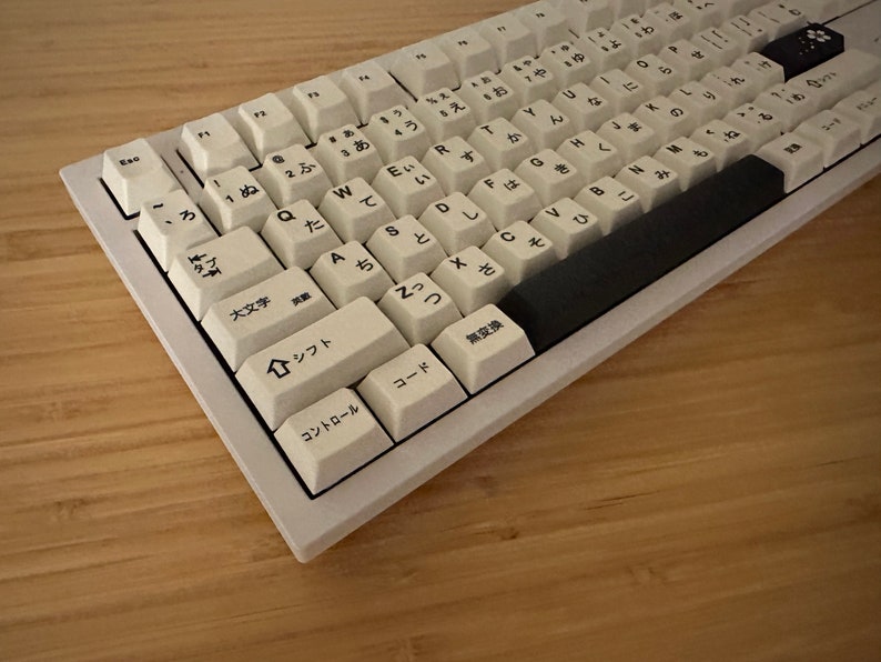 K&ouml;nnte beinhalten: Eine wei&szlig;e mechanische Tastatur mit japanischen Zeichen auf den Tasten. Die Tastatur hat eine schwarze Leertaste und eine schwarze Escape-Taste. Die Tastatur befindet sich auf einer Holzoberfl&auml;che.