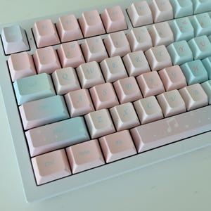May include: A white mechanical keyboard with pastel pink and blue keycaps. The keyboard has a standard layout with a number row, function keys, and arrow keys. The keycaps have a light, faded look, giving the keyboard a soft and dreamy aesthetic.