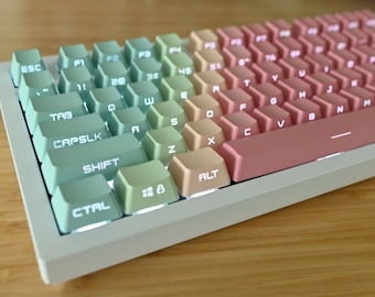 Op de afbeelding: Een wit mechanisch toetsenbord met pastelgroene, roze en beige toetsen. De toetsen zijn gelabeld met letters, cijfers en symbolen. Het toetsenbord heeft een wit frame en een witte basis.