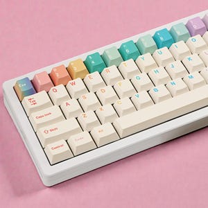 May include: A white mechanical keyboard with pastel-coloured keycaps. The top row of number keys features a gradient of colours, from pink to purple. The alphanumeric keys are off-white with colourful lettering. The keyboard sits on a pink surface.