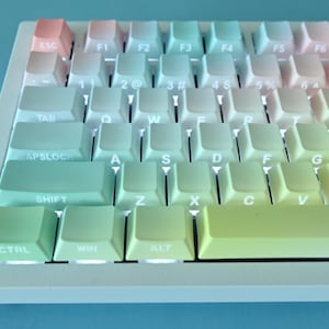 May include: A mechanical computer keyboard featuring a gradient colour scheme on the keycaps, transitioning from soft pink and light blue to pale yellow and green. The keys have white lettering, and the keyboard frame is a neutral off-white colour.
