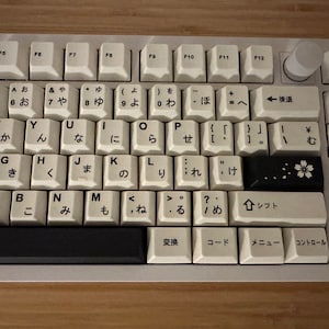 K&ouml;nnte beinhalten: Eine wei&szlig;e Tastatur mit schwarzen Tasten und japanischen Zeichen. Die Tastatur hat eine schwarze Leertaste und eine schwarze Taste mit einem wei&szlig;en Blumendesign.