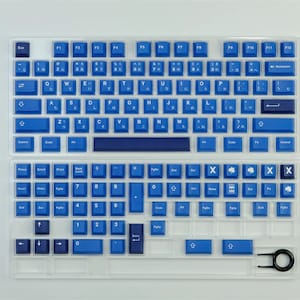 K&ouml;nnte beinhalten: Eine geteilte mechanische Tastatur mit blauen Tastenkappen und japanischen Schriftzeichen. Die Tastenkappen sind in einem leuchtenden Blau gehalten, mit wei&szlig;er Beschriftung. Die Tastatur befindet sich in einem transparenten Kunststoffgeh&auml;use, und ein Keycap-Abzieher ist enthalten.