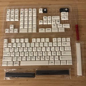 K&ouml;nnte beinhalten: Eine wei&szlig;e mechanische Tastatur mit japanischen Tastenkappen. Die Tastatur befindet sich in einem durchsichtigen Kunststoffgeh&auml;use und enth&auml;lt einen Satz schwarzer und wei&szlig;er Tastenkappen, einen wei&szlig;en Tastenkappenzieher und zwei schwarze Stabilisatoren.