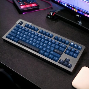 K&ouml;nnte beinhalten: Eine mechanische Tastatur mit einem dunkelgrauen Rahmen und marineblauen Tastenkappen. Die Tastatur hat ein Standardlayout mit alphanumerischen Tasten, Funktionstasten und einem Nummernblock. Die Tastenkappen haben wei&szlig;e Buchstaben und Symbole. Die Tastatur befindet sich auf einer dunklen Oberfl&auml;che.