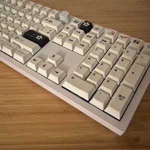 K&ouml;nnte beinhalten: Eine wei&szlig;e mechanische Tastatur mit schwarzen Tastenkappen und japanischen Zeichen. Die Tastatur verf&uuml;gt &uuml;ber einen Nummernblock und Pfeiltasten. Die Tasten sind mit japanischen Zeichen und englischen Buchstaben beschriftet.