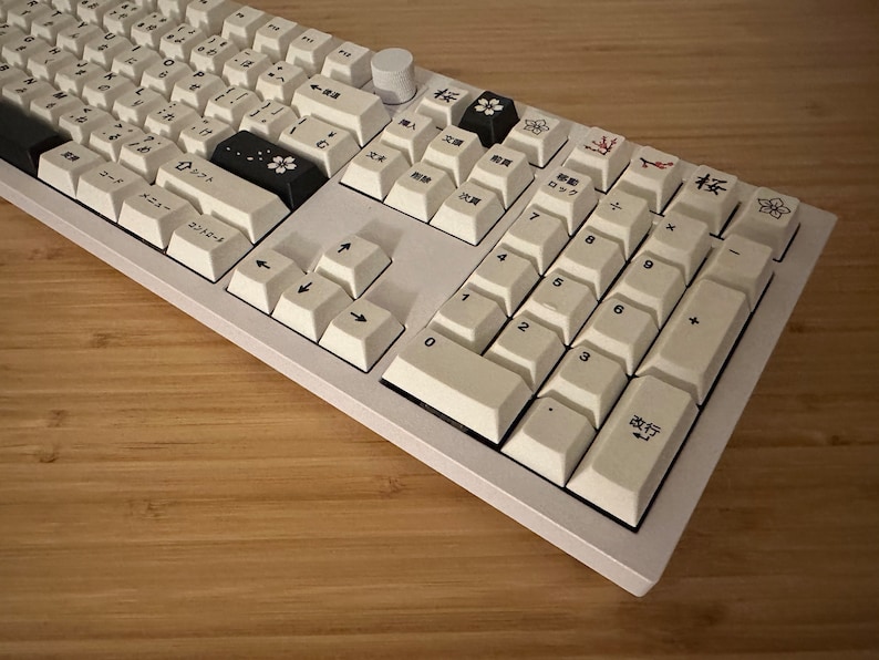 K&ouml;nnte beinhalten: Eine wei&szlig;e mechanische Tastatur mit schwarzen Tastenkappen und japanischen Zeichen. Die Tastatur verf&uuml;gt &uuml;ber einen Nummernblock und Pfeiltasten. Die Tasten sind mit japanischen Zeichen und englischen Buchstaben beschriftet.