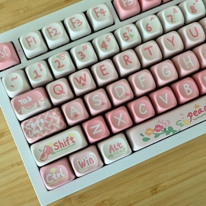 May include: A white keyboard with pink keys and a floral design on the space bar. The keys have various symbols and letters printed on them, including "Esc", "Tab", "Caps Lock", "Shift", "Ctrl", "Win", "Alt", and "& Peach +00".
