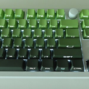 以下が含まれることがあります： キーに白い文字が書かれた緑色のメカニカルキーボード。キーボードは右側にテンキー、上部にファンクションキーの列があります。