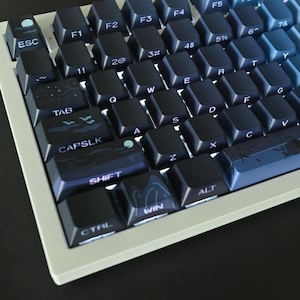 Könnte beinhalten: Eine weiße und schwarze mechanische Tastatur mit blauen Tastenkappen. Die Tastenkappen zeigen verschiedene Symbole und Designs, darunter einen Mond, Sterne, Vögel und Berge. Die Tastatur hat einen weißen Rahmen und eine schwarze Basis.